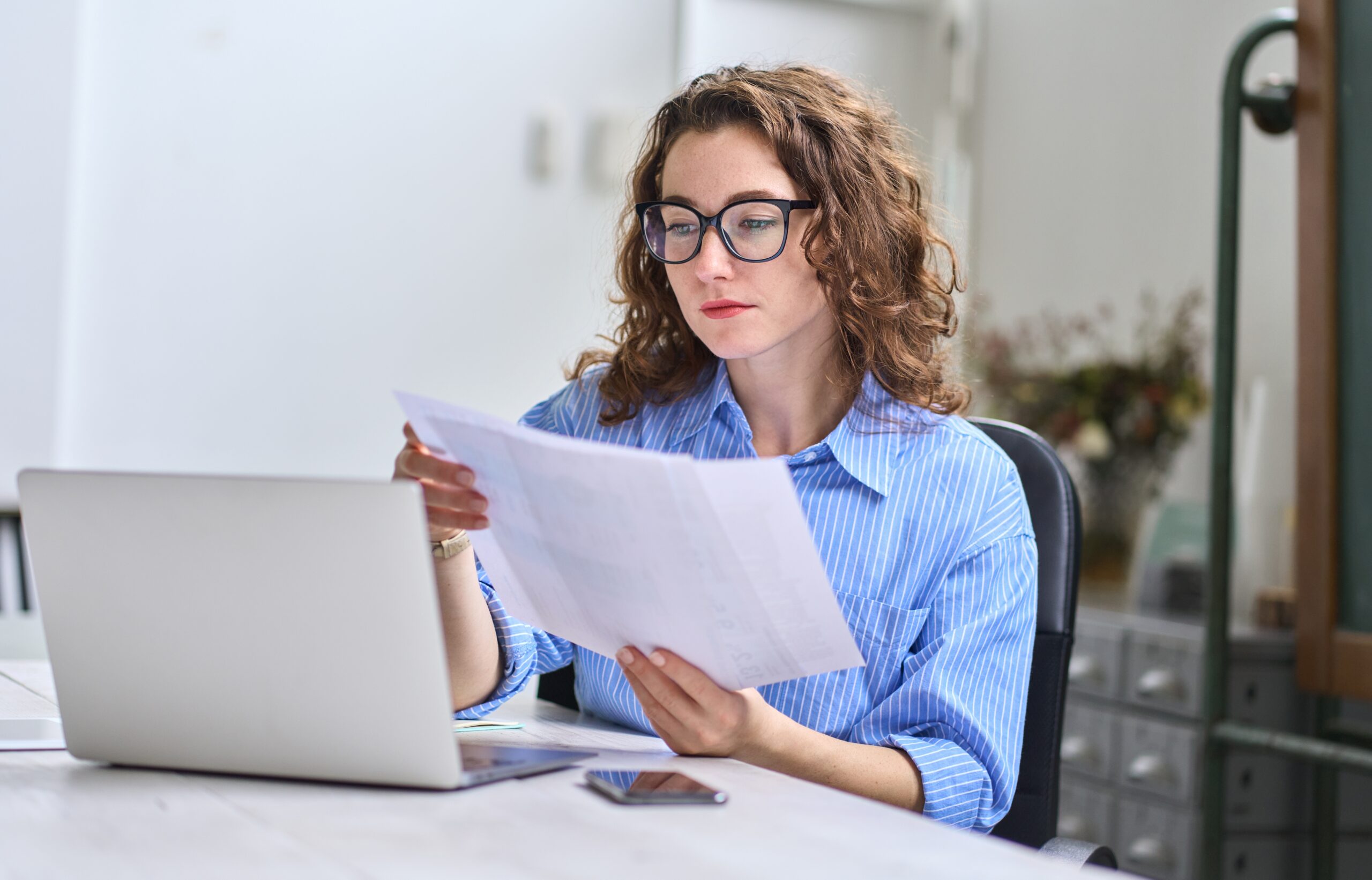 woman wearing glasses reviewing budget documents in front of her laptop computer